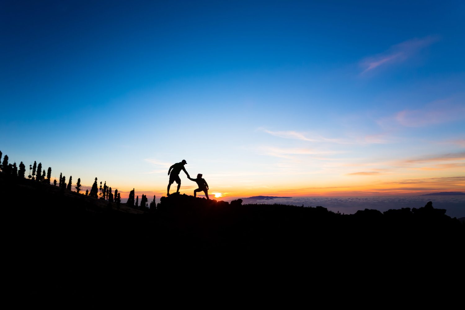 Teamwork couple helping hand trust help silhouette in mountains, sunset. Team of climbers man and woman hikers, help each other on top of mountain, beautiful inspirational sunset landscape on Tenerife Canary Islands